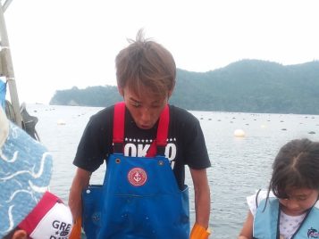 A man wearing blue waders stands on a boat between two school-aged children. The photograph captures a fish leaping from a bucket and the fisherman's surprised expression.